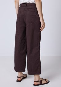 Pantalons évasés marron avec une taille élastique, deux poches arrière et un tissu légèrement texturé, associés à des sandales noires.