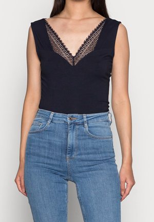 Frau trägt ein ärmelloses schwarzes Top mit Spitzen-V-Ausschnitt, das in hoch taillierte blaue Jeans gesteckt ist, vor weißem Hintergrund.