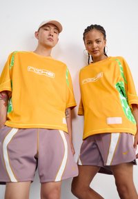 Tricouri oversized portocalii cu accente verzi, asortate cu pantaloni scurți violet deschis cu dungi albe și logo-ul Nike, pe modele care stau unul lângă celălalt.