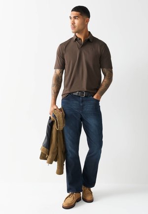 Mężczyzna z tatuażami, ubrany w brązową koszulkę polo i niebieskie dżinsy, trzymający brązową kurtkę w jednej ręce, stojący na jednolitym białym tle.