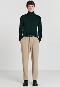 Dunkelgrüner Rollkragenpullover mit langen Ärmeln, kombiniert mit beigefarbenen, faltigen Hemdhosen; getragen mit einem schwarzen Gürtel und schwarzen Stiefeletten.