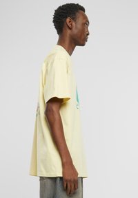 Yellow cotton t-shirt with short sleeves, relaxed fit, featuring a graphic design on the front in teal and pink, worn with gray pants.