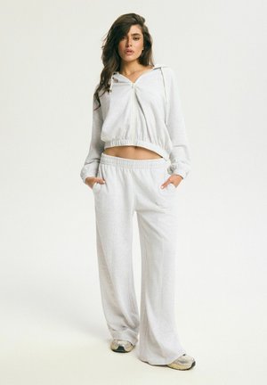 Žena stojaca s rukami v vreckách, oblečená v svetlosivej kapucňovej skrátenej mikine na zips, k nej ladia široké teplákové nohavice a béžové tenisky.