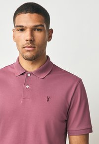 Polokošeľa v farbe fialovej s klasickým golierom, dvoma gombíkmi a malým vyšívaným logom na hrudi. Tkanina pôsobí textúrovaným a priedušným dojmom.