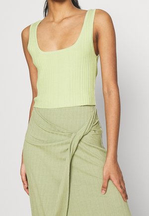 Vrouw draagt een lichtgroene, geribbelde mouwloze top en bijpassende wikkelrok, getoond van nek tot midden-dij tegen een effen achtergrond.