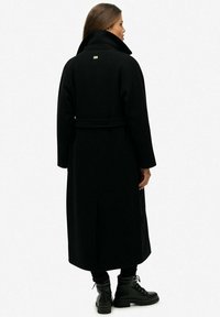 Sort uldblandings trenchcoat med høj krave, bælte i taljen og sidelommer. Har en metal logo detaljer på ryggen.