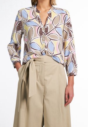 Vrouw draagt een pastelblouse met een abstract bladpatroon, ingestopt in beige, hoge taille, wijde broek met stoffen riem gestrikt in de taille.