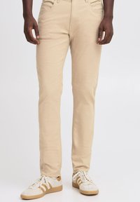 Pantaloni beige realizzati in tessuto misto cotone, con una vestibilità slim, tasche standard e una superficie liscia. Abbinati a scarpe da ginnastica bianche.
