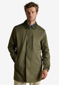 Vīrietis, kurš valkā olīvu zaļu līdz ceļiem garu apmetni ar tumšu apkakli un kabatām, apvienojot to ar gaišiem biksēm, stāv pret vienkāršu fona.