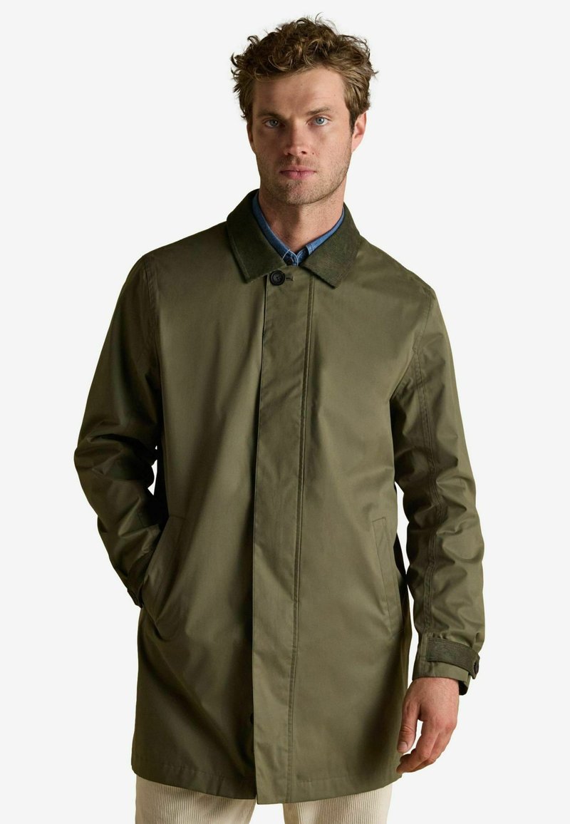 Vīrietis, kurš valkā olīvu zaļu līdz ceļiem garu apmetni ar tumšu apkakli un kabatām, apvienojot to ar gaišiem biksēm, stāv pret vienkāršu fona.