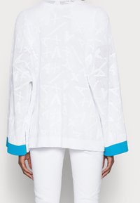 Bílý pletený svetr s geometrickým vzorem, s dlouhými rukávy a tyrkysovým žebrovaným lemem. Design s otevřeným zadním dílem a prodyšná textura.