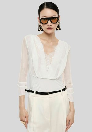 Donna che indossa una camicetta bianca trasparente con dettagli in pizzo, pantaloni color crema a vita alta, cintura nera, grandi orecchini dorati e occhiali da sole oversize con lenti colorate.
