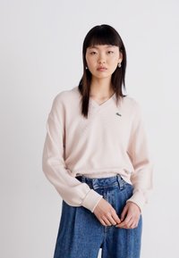 Pull V-neck texturé rose clair à manches longues, associé à un jean en denim bleu taille haute. Petit logo vert sur la poitrine gauche.