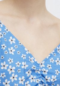 Světle modré šaty s V-výstřihem, zdobené bílými květinami a černými akcenty. Tkanina vypadá strukturovaně s jemným leskem.