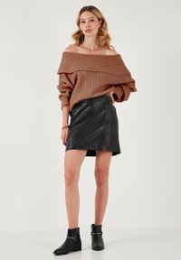 Suéter marrón de canalé con off-the-shoulder, mini falda de imitación cuero negra con abertura frontal, combinada con botas negras hasta el tobillo con acentos metálicos.