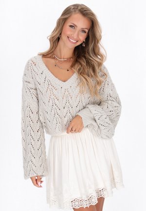 Maglione grigio chiaro lavorato a maglia con scollo a V e maniche ampie, abbinato a una gonna bianca con orlo in pizzo.