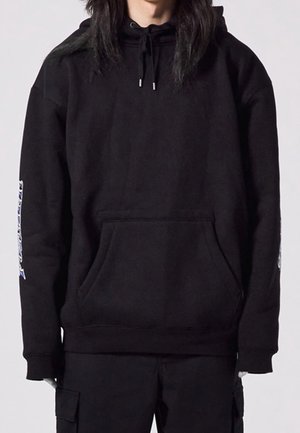 Person iført en sort hættetrøje med kængurulomme og snor i hætten, kombineret med sorte cargobukser mod en neutral baggrund.