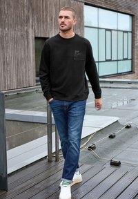 Černá mikina s kulatým výstřihem, zdobená šedým grafickým potiskem na přední straně; v kombinaci s modrými džíny a bílými teniskami s zelenými akcenty.