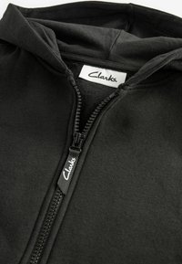 Černá mikina na zip vyrobená z měkké tkaniny s kapucí. Obsahuje štítek s logem Clarks a černý zip. Hladká textura.