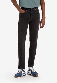 Svarta denimjeans med slim fit, femficksdesign, kontrasterande sömmar och kopparfärgade detaljer, kombinerade med blåa sportskor.