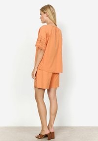 Completo in cotone arancione composto da due pezzi, con una maglia ampia e maniche con finiture in pizzo, abbinato a pantaloni corti, mostrato di spalle. Sandali con tacco a blocco marroni.