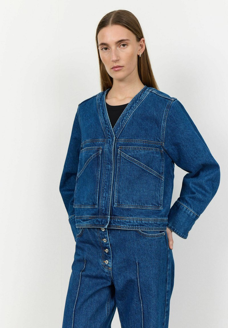 Modra jeans jakna z V-izrezom, dvema sprednjima žepoma in kontrastnim šivanjem. Ima ohlapen kroj in manšete z rahlim razširitvami.