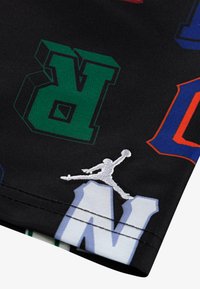 Pantalones cortos de tela negra con letras en bloque de colores (verde, rojo, azul) y un logotipo de Jumpman bordado en blanco. Textura suave, diseño deportivo.