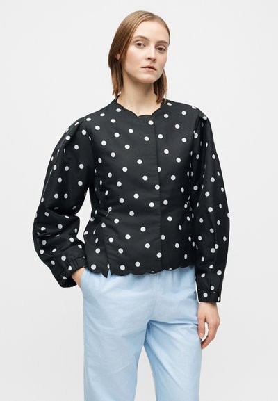 Juoda striukė su baltomis taškelėmis, suapvalintu iškirpimu, pūstomis rankovėmis ir bangelėmis į apačią. Audinys atrodo lygus ir lengvas, derinamas su šviesiai mėlynomis kelnėmis.