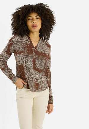 Femme aux cheveux bouclés portant une chemise à manches longues marron à motifs et un pantalon beige clair, debout avec une main dans la poche devant un fond blanc.