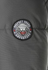 Tela acolchada gris con un parche circular bordado que presenta montañas, "RESPECT BRAND," "ADVENTURE," y el año "1997" en rojo y blanco.