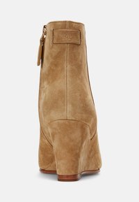 Stivale alla caviglia in suede beige con tacco a zeppa, dotato di una cerniera posteriore e un'etichetta rettangolare sul retro. Texture liscia con silhouette elegante.