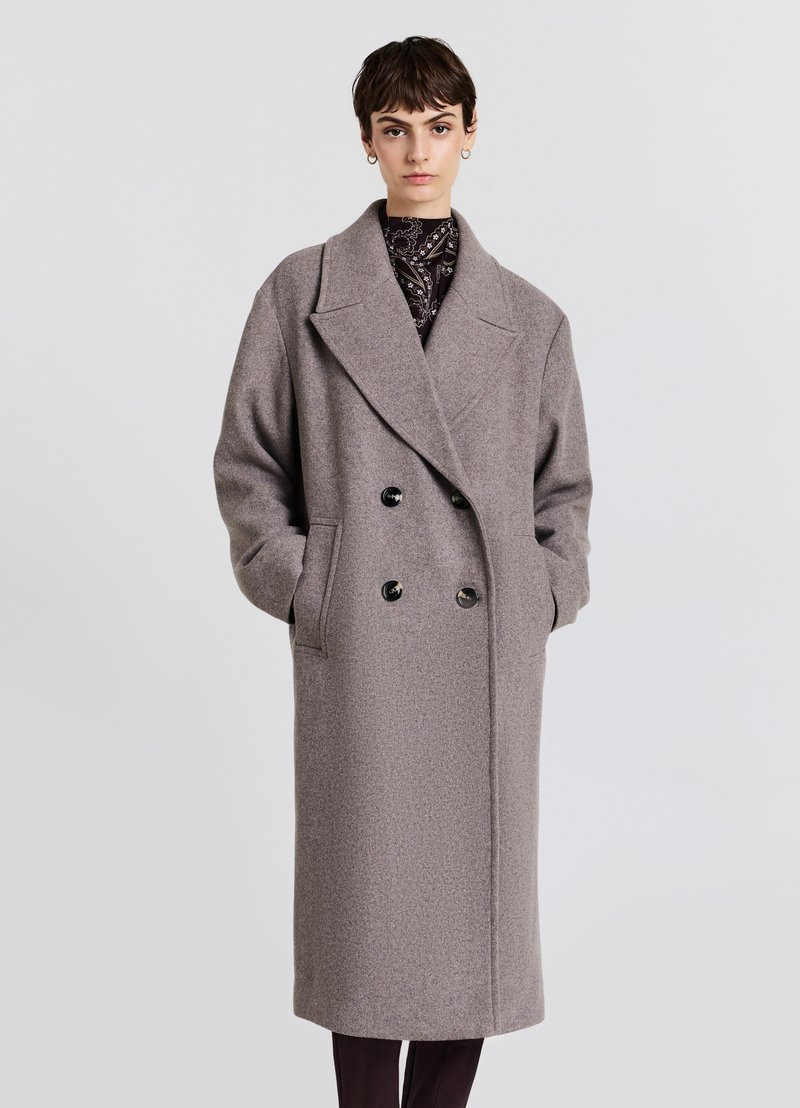 Cappotto doppiopetto in lana grigia con grandi rever, due tasche frontali e bottoni neri. Senza fodera, con vestibilità comoda e lunghezza fino a metà polpaccio.