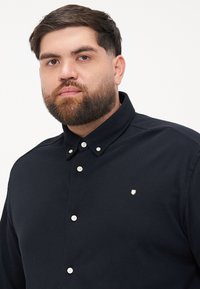Tummansininen pitkähihainen paita, jossa on napitettava kaulus, valkoiset napit ja pieni logo vasemmassa rinnassa. Sujuva kankaan rakenne.