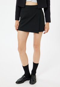 Schwarzer Wickelrock mit passendem Bund, glattem Stoff und minimalistischem Design. Kombiniert mit schwarzen Socken und glänzenden Schuhen.