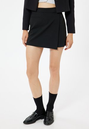 Schwarzer Wickelrock mit passendem Bund, glattem Stoff und minimalistischem Design. Kombiniert mit schwarzen Socken und glänzenden Schuhen.