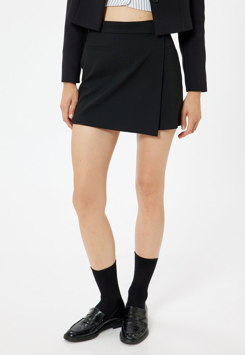 Schwarzer Wickelrock mit passendem Bund, glattem Stoff und minimalistischem Design. Kombiniert mit schwarzen Socken und glänzenden Schuhen.
