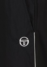 Schwarzes, leichtes Stoffkleidungsstück mit einem weißen, gestickten Logo unten links und kontrastierenden weißen Nähten entlang der Kanten.