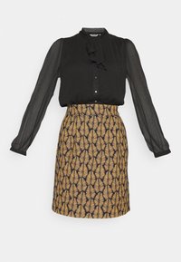 Blouse noire à manches longues en voile transparent et col noué, associée à une jupe mi-longue ornée d'un motif de feuilles dorées sur fond sombre.