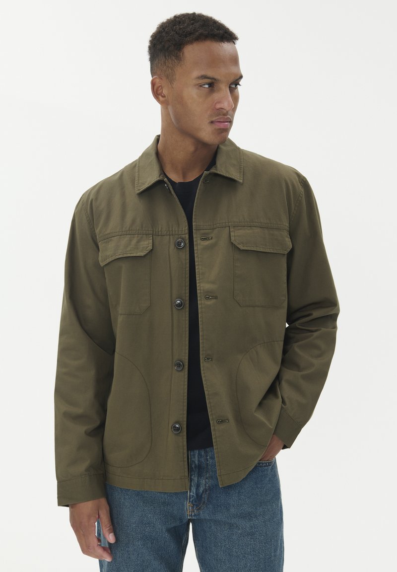 Olive zelena jakna iz teksturiranega blaga, s sprednjim gumbom, dvema žepoma na prsih in sproščenim krojem. Nosena čez temno srajco.