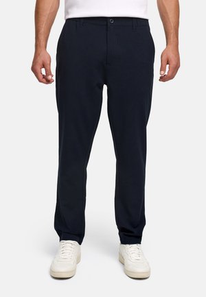 Homme portant un pantalon bleu marine ajusté et des baskets blanches, debout devant un fond clair uni.