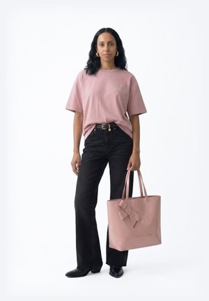 Mujer con cabello largo y negro, viste una camiseta rosa y jeans negros, sosteniendo un gran bolso rosa con un detalle de lazo.