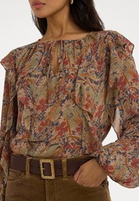 Blommig blus i genomskinligt tyg med volanger, med en nyckelhålsutskärning. Jordnära nyanser av grönt, rött och blått. Kombinerad med ett brunt bälte.