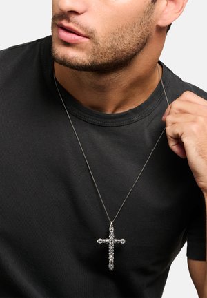 Silbernes Kreuzanhänger-Halsketten mit aufwendigen Details, an einer feinen Kette getragen, über einem schwarzen Rundhals-T-Shirt.
