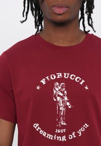 Burgundy cotton T-shirt with a round neckline featuring a white graphic of two figures and text, "Fiorucci dreaming of you," dated 1987.