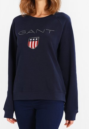 Kobieta nosząca granatową bluzę z logo GANT i czerwono-biało-niebieskim emblem emblem tarczy na piersi, w połączeniu z ciemnymi jeansami.
