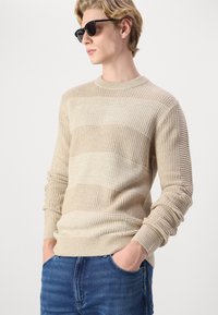 Maglione beige a maglia con strisce testurizzate, scollo tondo e polsini a costine. Indossato con jeans blu, mani in tasca e occhiali da sole neri.