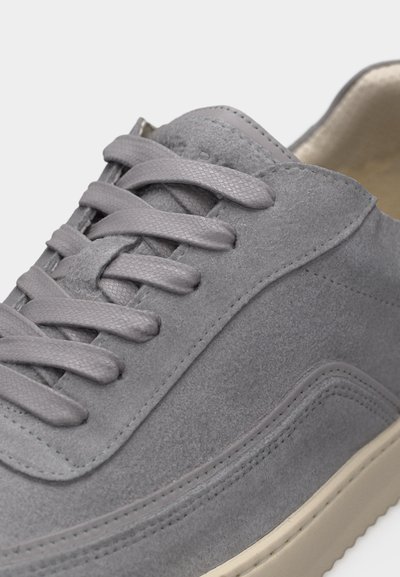 Gros plan sur une sneaker en daim gris avec des lacets texturés et une semelle couleur crème, mettant en valeur les coutures et la matière douce.