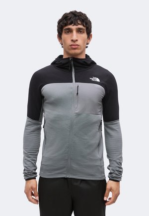 Młody mężczyzna w czarno-szarej kurtce na zamek błyskawiczny z kapturem oraz logo The North Face na piersi, stojący na jednolitym tle.