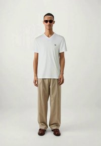 Λευκή μπλούζα με V λαιμόκοψη και μικρό λογότυπο στο στήθος, συνδυασμένη με μπεζ παντελόνια με φαρδιά πόδια και καφέ λοφότες, αξεσουάρ με σκούρα γυαλιά ηλίου.