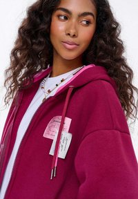 Bordový mikina s kapucí a růžovým interiérem a stahovacími šňůrkami, na přední straně s textovými štítky. Tkanina se zdá být měkká a strukturovaná.